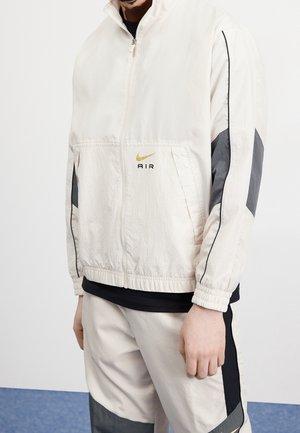 Mężczyzna ubrany w jasnobeżową kurtkę Nike Air zapinaną na zamek z czarnymi i szarymi akcentami na rękawach oraz pasujące spodnie, stojący na białym tle.