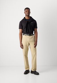 Zwarte shirt met korte mouwen met logo, beige broek en zwarte schoenen. Een trui om de nek voegt textuur en contrast toe aan de outfit.