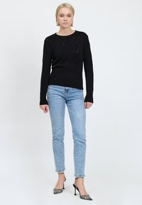Donna bionda in piedi contro uno sfondo bianco, vestita con un maglione nero lavorato a maglia, jeans skinny azzurri chiari e sandali neri con tacco alto e cinturini alla caviglia.
