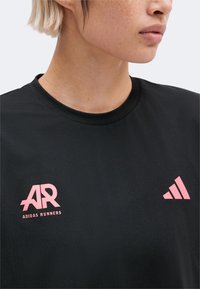 Κοντινό πλάνο ενός ατόμου που φοράει μαύρο T-shirt Adidas Runners με ροζ λογότυπα στο στήθος, κοντά ξανθά μαλλιά και ουδέτερο φόντο.