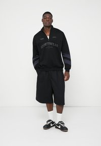 Pulover sport cu fermoar la gât de culoare neagră, cu textul "SPORTSWEAR", pantaloni scurți negri asortați și adidași negri cu accente albe, pe un fundal simplu.