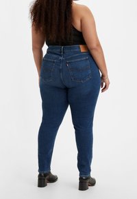 Modré džíny s vysokým pasem, hladkou texturou, pěti kapsami a hnědou koženou nášivkou na zadním pasu.