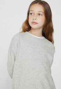 Šedá mikina s jemnou texturou, s kulatým výstřihem a volným střihem, která je vrstvená přes bílou košili na límci.