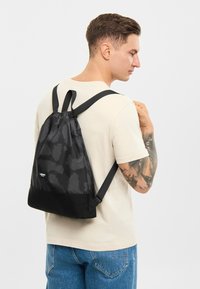 Rucsac negru cu șnur, cu o finisare mată texturată, are un model subtil de camuflaj, un fund negru și bretele ajustabile.