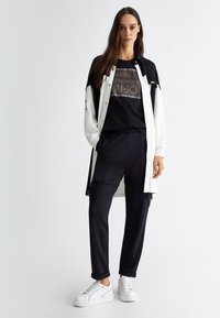 T-shirt nera con stampa grafica, giacca bianca a bottoni, pantaloni jogger neri e sneakers bianche. Tessuto morbido, vestibilità comoda e pantaloni con bordi risvolti.