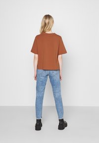 Kobieta z długimi blond włosami, nosząca luźną brązową koszulkę, jasnoniebieskie dżinsy i czarne buty na platformie, stoi zwrócona w stronę białej ściany.