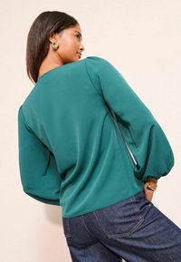 Teal blouse with balloon sleeves and a relaxed fit, paired with dark denim jeans. Features smooth texture and simple neckline design.