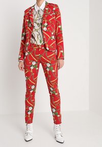 Röd blommönstrad kavaj och byxor med guldkedjor. En vit skjorta med krage under. Stylad med vita anklarstövlar.