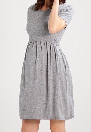 Femme portant une robe grise à manches courtes, longueur au genou, avec une taille cintrée et une jupe froncée, sur un fond clair uni.