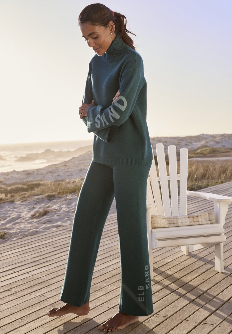 Türkiser Strickpullover mit Rollkragen und dem Schriftzug "STAND" am Ärmel, kombiniert mit weit geschnittenen Hosen, die seitlich "ELB SAND" zeigen, barfuß getragen.