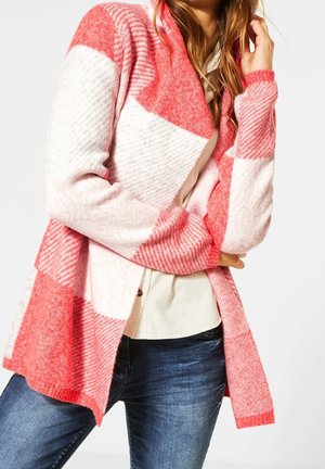 Femme portant un cardigan ouvert à carreaux rouges et blancs sur un chemisier crème et un jean bleu, reposant sa main près du visage.