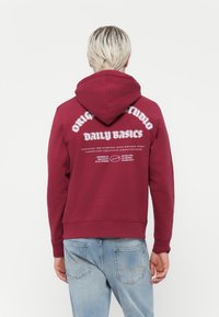 Sweat à capuche bordeaux avec un cordon de serrage, présentant un texte blanc au dos. Fabriqué en tissu texturé, avec des poignets et un ourlet côtelés.