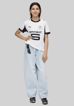 Giovane in piedi con la mano sui fianchi, che indossa una maglietta sportiva bianca e nera, jeans azzurri a gamba larga e sneakers nere.