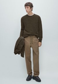 Brązowy miękki sweter dzianinowy, beżowe zwężane spodnie ze ściągaczem oraz ciemne buty. Trzyma zieloną kurtkę. Prosty, swobodny strój.