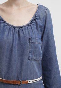 Nainen, jolla on yllään sininen farkkupaita pitkällä hihalla, jossa on rintatasku, sekä ruskea nahkavyö, jossa on valkoista palmikkokuosia.