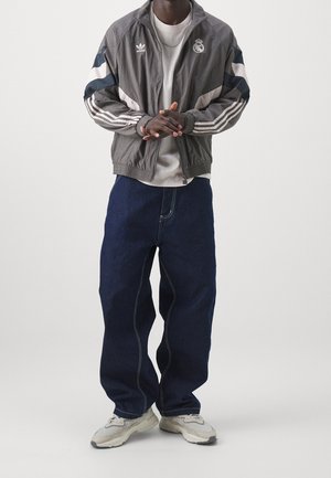 Mand iført grå Adidas og Real Madrid træningsjakke, hvid skjorte, løstsiddende mørkeblå jeans og hvide sneakers, stående med hænderne foldet.