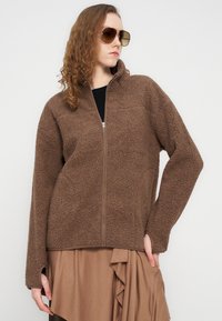 Καφέ μπουφάν φλις με ψηλό γιακά και φερμουάρ στο μπροστινό μέρος. Διαθέτει υφή υφάσματος και άνετη εφαρμογή με θηλάκια για τους αντίχειρες στα μανίκια.