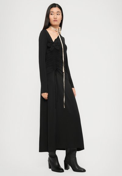 Hosszú, fekete ruha redőzött mellrésszel, hosszú ujjakkal és húzózsinóros részlettel. Fémes arany szegély található a nyak körül, valamint fekete magassarkú csizmák.