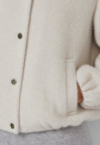 Lichtbeige textuurtrui met metalen drukknoopjes, voorzien van een zijzak en gathered manchetten voor een snug fit. Zacht, comfortabel materiaal.
