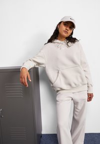 Šviesiai smėlinis džemperis su priekinėmis kišenėmis ir raišteliu, derinamas su atitinkančiomis kelnėmis. Pavyzdys turi mažą baltą Nike logotipą. Fone matyti pilkas spintelė.
