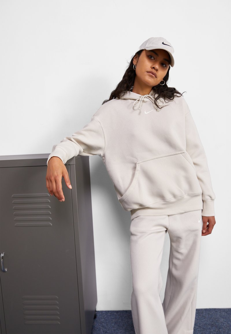 Šviesiai smėlinis džemperis su priekinėmis kišenėmis ir raišteliu, derinamas su atitinkančiomis kelnėmis. Pavyzdys turi mažą baltą Nike logotipą. Fone matyti pilkas spintelė.