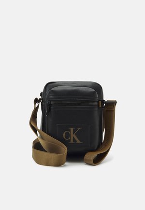 Musta nahkainen olkalaukku vetoketjullisella sulkemisella, jossa on ruskea olkahihna ja etuosassa kohokuvioitu "CK"-logo.