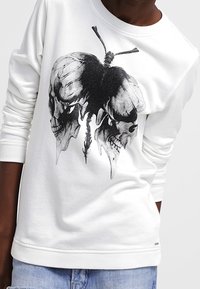 Vit sweatshirt med en grafisk design av överlappande dödskallar och en fjäril. Tillverkad av mjukt material med ribbade ärmslut och fåll.