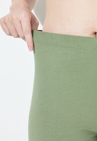 Ręka odciągająca pas zielonych, rozciągliwych spodni od gołej talii i brzucha, ukazując dopasowanie i elastyczność.