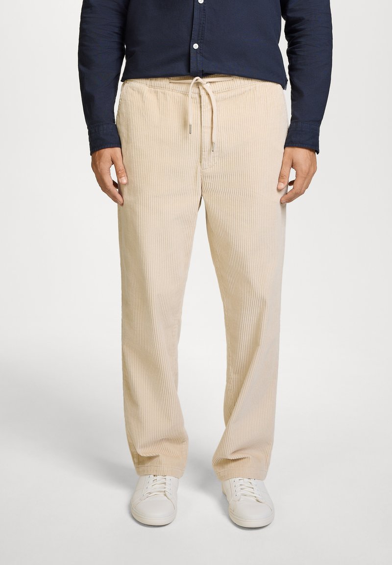 Pantalones de pana beige con costuras verticales, cintura con cordón y bolsillos laterales, combinados con zapatillas blancas. Adecuados para un estilo casual.