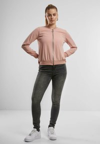 Světle růžová zipová bunda s vysokým límcem, přiléhavý design, žebrované manžety a spodní lem, nošená přes tmavě šedé džíny a bílé tenisky.