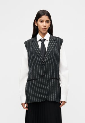 Mujer con camisa blanca y corbata negra, debajo de un chaleco negro sin mangas con rayas finas, combinada con una falda plisada negra, sobre un fondo blanco.