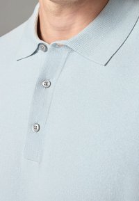 Světle modré pletené polo tričko s plochým límečkem, třemi knoflíky a hladkou texturou.