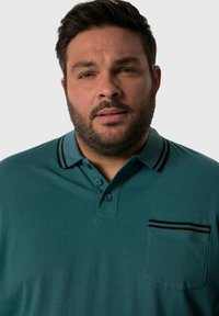 Polo shirt turchese realizzato in tessuto morbido, con colletto a righe bianche e nere, apertura con tre bottoni e taschino sul petto con righe coordinate.
