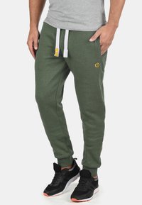 Green sweatpants made of soft fabric, featuring a relaxed fit, white drawstring, and a small yellow logo on the thigh.