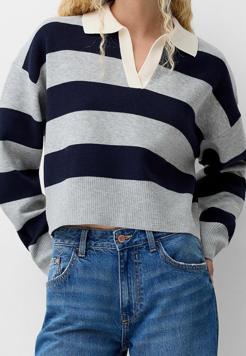 Personne portant un pull à larges rayures bleu marine et gris avec un col blanc et un jean bleu, mains dans les poches, montrée du cou aux hanches.
