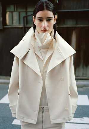 Vrouw met stijl donker haar draagt een oversized beige jas en een shirt met hoge hals, staand buiten op een straat met een vage achtergrond.
