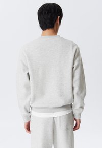 Person wearing light gray sweatshirt and matching sweatpants, standing facing away against a plain white background.