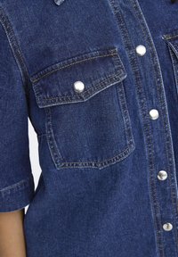 Denim shirt in deep blue with silver snap fasteners and two patch pockets, featuring short sleeves and contrasting yellow stitching.