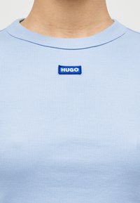 Ανοιχτό μπλε ριμπ κοντομάνικο μπλουζάκι με στρογγυλή λαιμόκοψη και μπλε ορθογώνιο ύφασμα με τη λέξη "HUGO" σε λευκό. Υφή υφάσματος, casual σχεδιασμός.