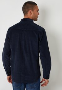 Marineblå kordfløjlsskjorte med lange ærmer, knapmanchetter og detaljer med plissering på ryggen. Glat overflade, afslappet pasform og klassisk kravedesign.