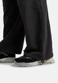Footwear features black suede and mesh upper, gray rubber sole with textured pattern, and Vibram branding on the side. Paired with black pants.