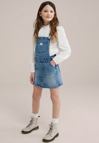 Denim overall obleka v svetlo modri barvi z nastavljivimi naramnicami, sprednjim žepom in prilegajočim dizajnom; združena z belo teksturirano srajco z dolgimi rokavi in bež škornji.