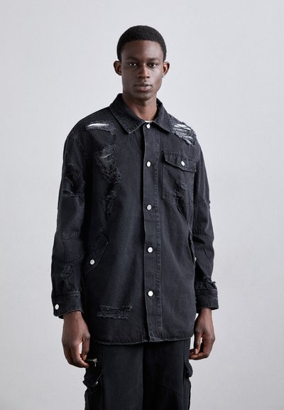 Black denim jacket featuring distressed patches, silver buttons, a classic collar, and two front pockets. Long sleeves with buttoned cuffs.