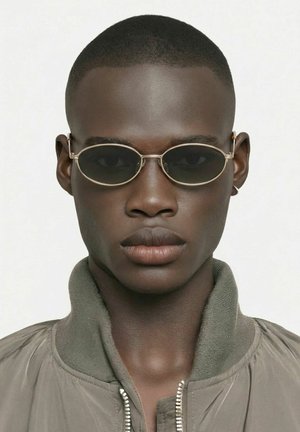 Jeune homme noir aux cheveux courts portant des lunettes de soleil ovales petites à monture dorée et une veste grise zippée sur un fond uni.