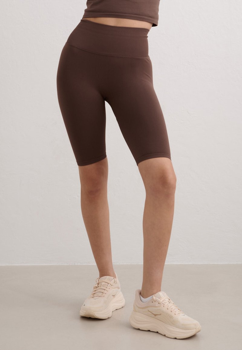 Personne portant un short de vélo ajusté taille haute marron foncé et des baskets beiges épaisses, debout sur un sol gris lisse contre un mur clair.