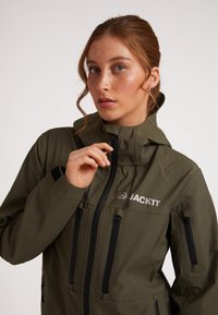 Giacca impermeabile verde oliva con dettagli neri. Chiusura a zip completa, cappuccio regolabile, molteplici tasche con zip e logo ben visibile sul petto.