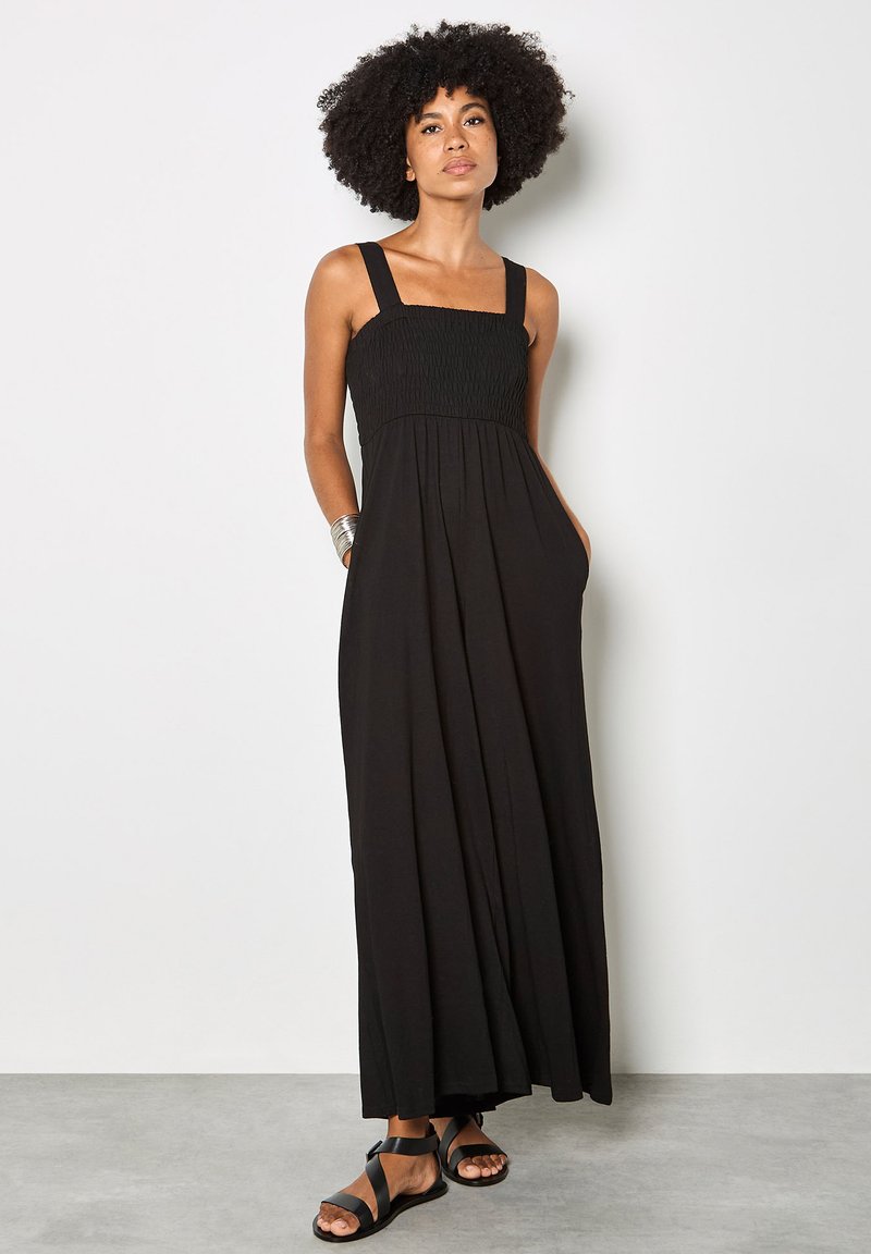 Femme aux cheveux bouclés portant une robe maxi noire sans manches et des sandales noires, debout avec les mains dans les poches de la robe contre un mur blanc.