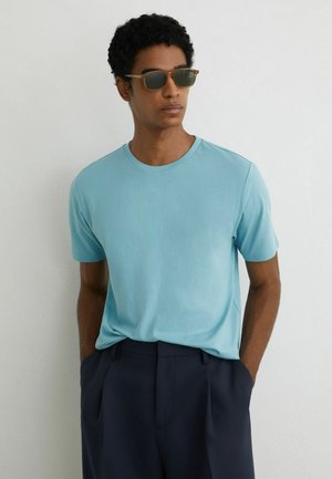 Ung mand iført en lyseblå t-shirt med korte ærmer, mørkeblå bukser og briller med skildpaddeskal, stående med hænderne i lommerne.