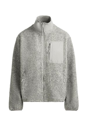 Jeune personne aux cheveux courts portant une casquette noire, une veste polaire gris clair avec poches zippées, une chemise blanche et un pantalon beige sur un fond uni.