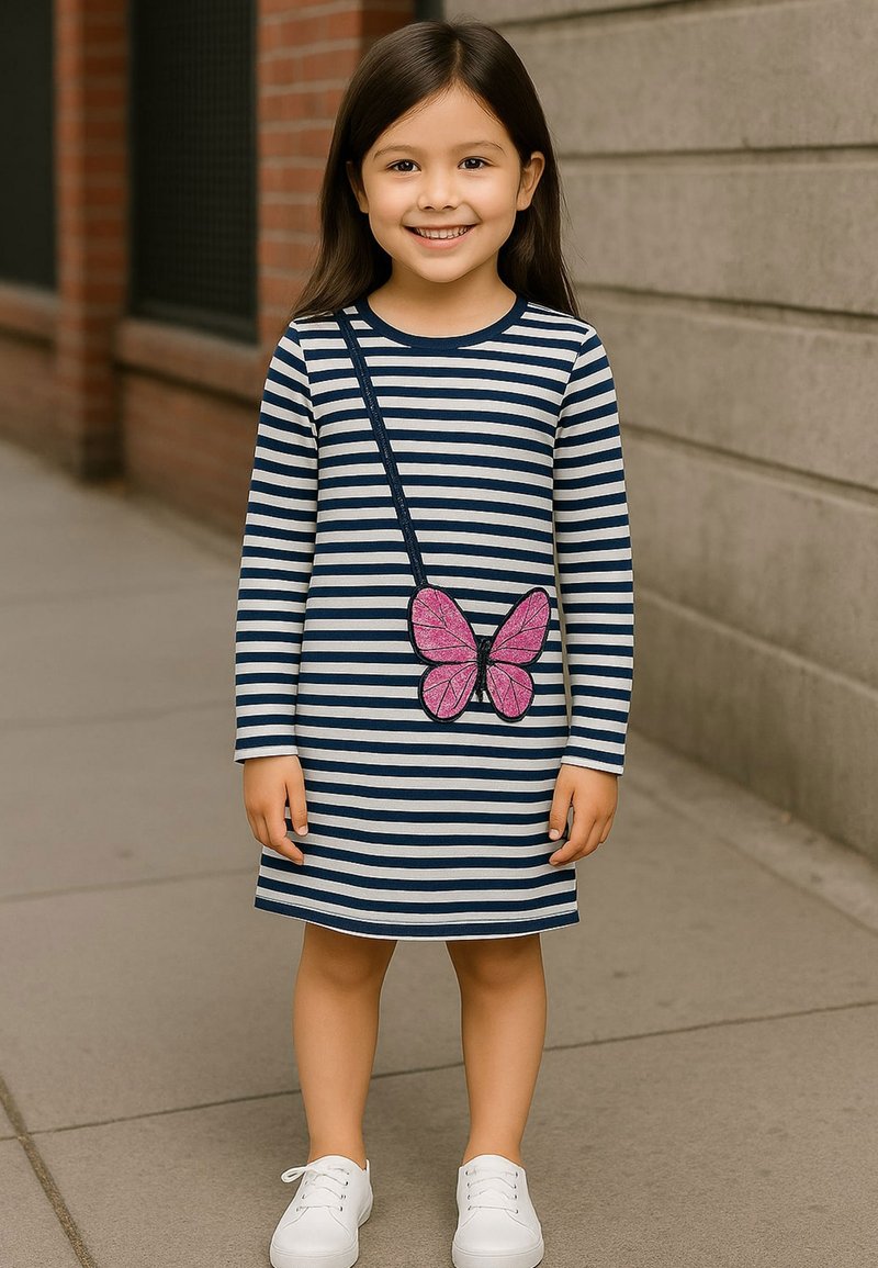Gestreiftes langärmliges Kleid in Marineblau und Weiß, mit einer rosa Schmetterlingsapplikation. Kombiniert mit weißen Sneakers, stehend auf einem Bürgersteig.